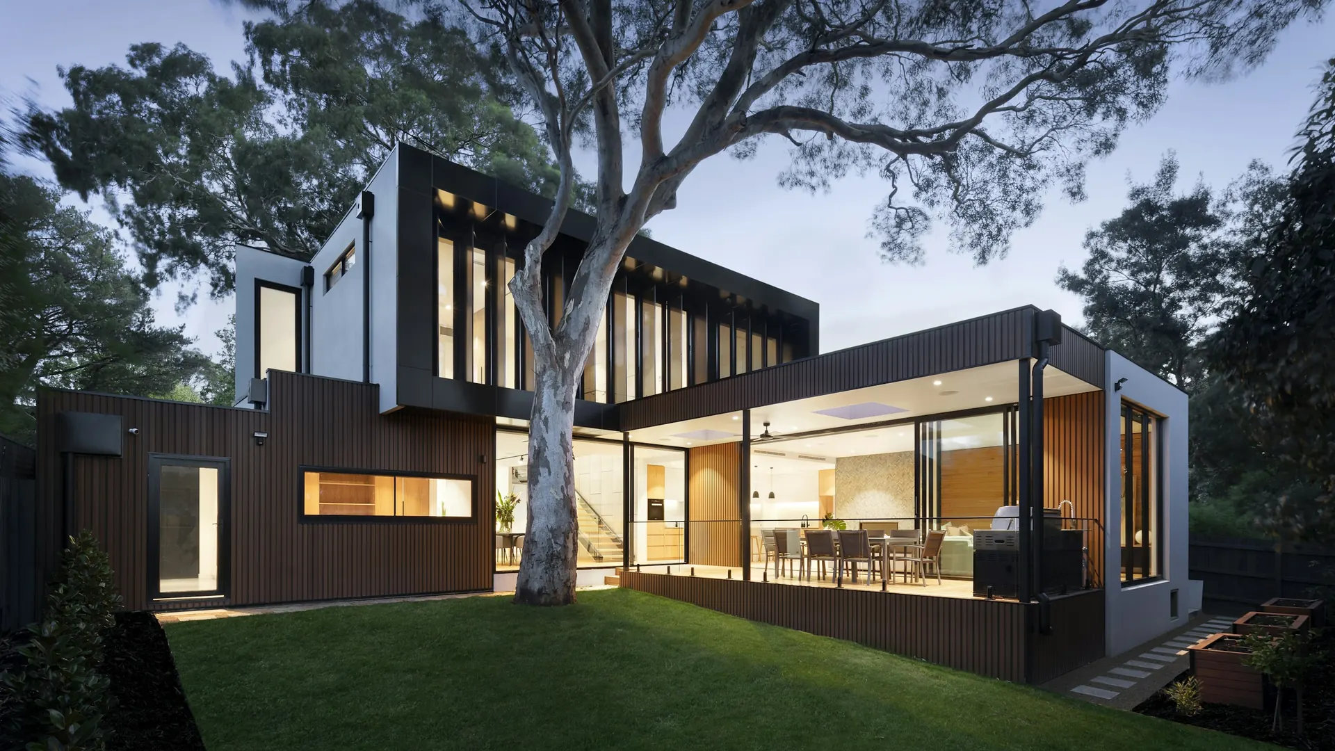Modern home exterior at dusk with lit interior, tree